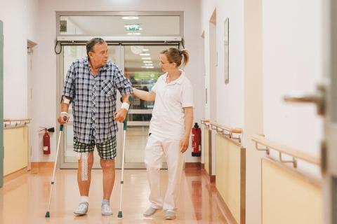 Ein orthopädischer Patient übt unter Anleitung einer Physiotherapeutin das Laufen an Gehhilfen auf dem Krankenhausflur.
