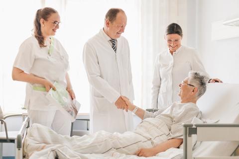 Zwei Ärzte und eine Krankenschwester am Bett einer älteren Patientin, ihr die Hand schüttelnd nach überstandener Operation.