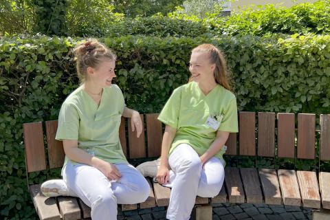 Freiwilligendienst im Krankenhaus: Lea (links) und Annemarie (rechts) leisten aktuell gerade ihr FSJ bzw. BFD-Jahr im St. Joseph-Stift Dresden. Hier zusammen auf einen Bank im Patientengarten, Krankenhausgebäude im Hintergrund.