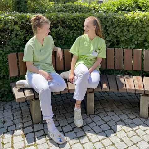 Freiwilligendienst im Krankenhaus: Lea (links) und Annemarie (rechts) leisten aktuell gerade ihr FSJ bzw. BFD-Jahr im St. Joseph-Stift Dresden. Hier zusammen auf einen Bank im Patientengarten, Krankenhausgebäude im Hintergrund. Größere Aufnahme.