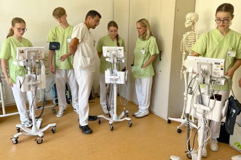 Vitalwerte am Patientenbett: Praxisanleiter und Freiwillige bei einer Unterweisung im Krankenhaus.