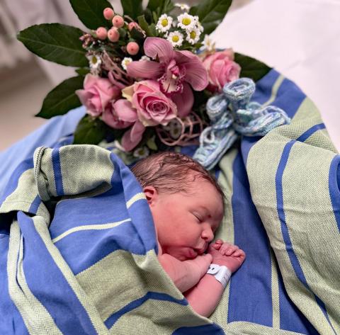 Neujahrsbaby Theo am 1.1.2025: Eingehüllt in eine Decke schläft der neugeborene Junge friedlich. In der Bildecke die Blumen und selbstgestrickten neuen Babyschuhe.