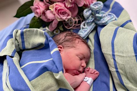 Neujahrsbaby Theo am 1.1.2025: Eingehüllt in eine Decke schläft der neugeborene Junge friedlich. In der Bildecke die Blumen und selbstgestrickten neuen Babyschuhe.