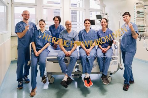 Team der ITS in einem Patientenzimmer auf dem Bett sitzend, mit Spruchband "Herzlich willkommen!"