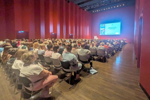 Blick in den Saal während eines Vortrages: volle Stuhlreihen, Podium und Redner vor der Videoleinwand.