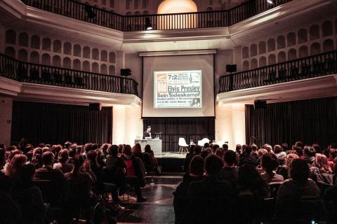 Szene während einer Lesung: ein abgedunkelter Saal vollbesetzt, auf dem Podium sitzt im Scheinwerferlicht Eric Wrede.