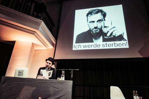 Blick in eine Lesung mit Eric Wrede: abgedunkelter Saal, Podium erleuchtet, Eric Wrede liest im Schein der Lampe aus einem Buch.