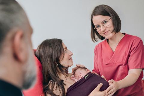Kampagnenmotiv Geburtshilfe im St. Joseph-Stift 2026: Geborgen und sicher im St. Joseph-Stift Dresden, mit 24/7 kinderärztlicher Betreuung. Abbildung zeigt Mutter und Baby mit Hebamme, im Anschnitt unscharf den Vater des Kindes. Headerbild für Teaserslide Geburtshilfe