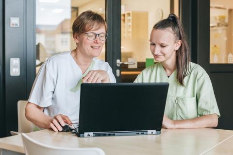 Pflegeausbildung in der Berufsschule: Berufsschullehrerin und Pflegeschülerin hinter einem Rechner auf der Station im Krankenhaus.