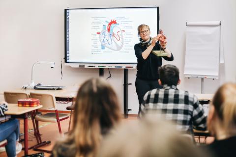 Schnupperstunde in der Medizinischen Berufsfachschule: Klassenraum mit Schülerinnen und Schülern, Lehrerin steht vorn und erklärt etwas am Modell, dahinter Flatscreen mit Unterrichtsmaterial.