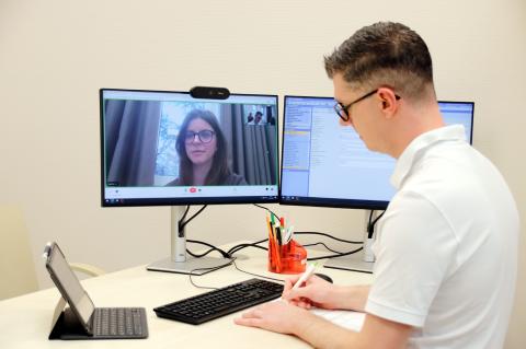 Videosprechstunde, exemplarisches Bild: Arzt in weißer Berufsbekleidung im Videocall mit Patientin, die auf dem Monitor zu sehen ist. Er macht sich während des Gesprächs Notizen.