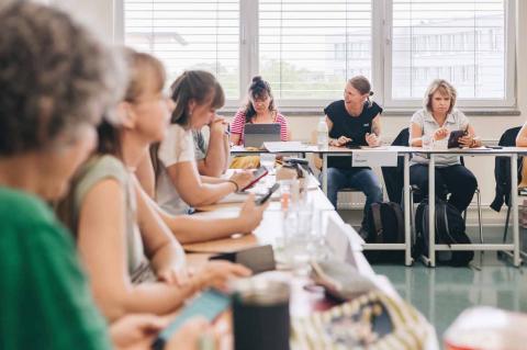 Akademie für Palliativmedizin und Hospizarbeit, Blick in einen laufenden Kurs, Teilnehmende auf Stühlen an Arbeitstischen während des Seminars im Gespräch untereinander.