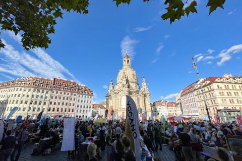 Gastmahl Dresden 2026 – Impressionen vom Neumarkt, im Hintergrund die Frauenkirche, im Vordergrund Tisch, bunte Schirme, Beachflags und massenhaft Menschen, buntes Treiben.