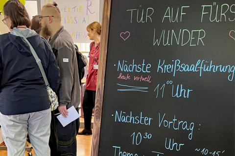 "Tür auf fürs Wunder" steht auf einer Kreidetafel im Eingangsbereich des Bildungszentrums am Krankenhaus St. Joseph-Stift Dresden, wo der erste Tag der offenen Tür der Geburtsklinik stattfand. Dahinter im Anschnitt Besucherinnen und Besucher der Veranstaltung, die sich an den Ständen im Foyer über Angebote informieren und Beratungen in Anspruch nehmen.