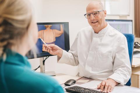 Plastische Chirurgie, Sprechstunde in der Zentralambulanz, Arzt frontal, mit dem Rücken zum Fotografen sitzt eine weibliche Patientin, auf dem Screen neben dem Arzt der Oberkörper einer Frau, der im Rahmen eines operativen Eingriffs plastisch-chirurgisch angepasst werden soll.