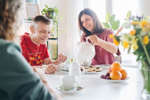 Im Tageshospiz, Kaffee und Kuchen am Kaffeetisch; Pflegerin gießt einem Gast Kaffee aus einer Kanne nach, es stehen Kuchen und Blumen auf dem Tisch.